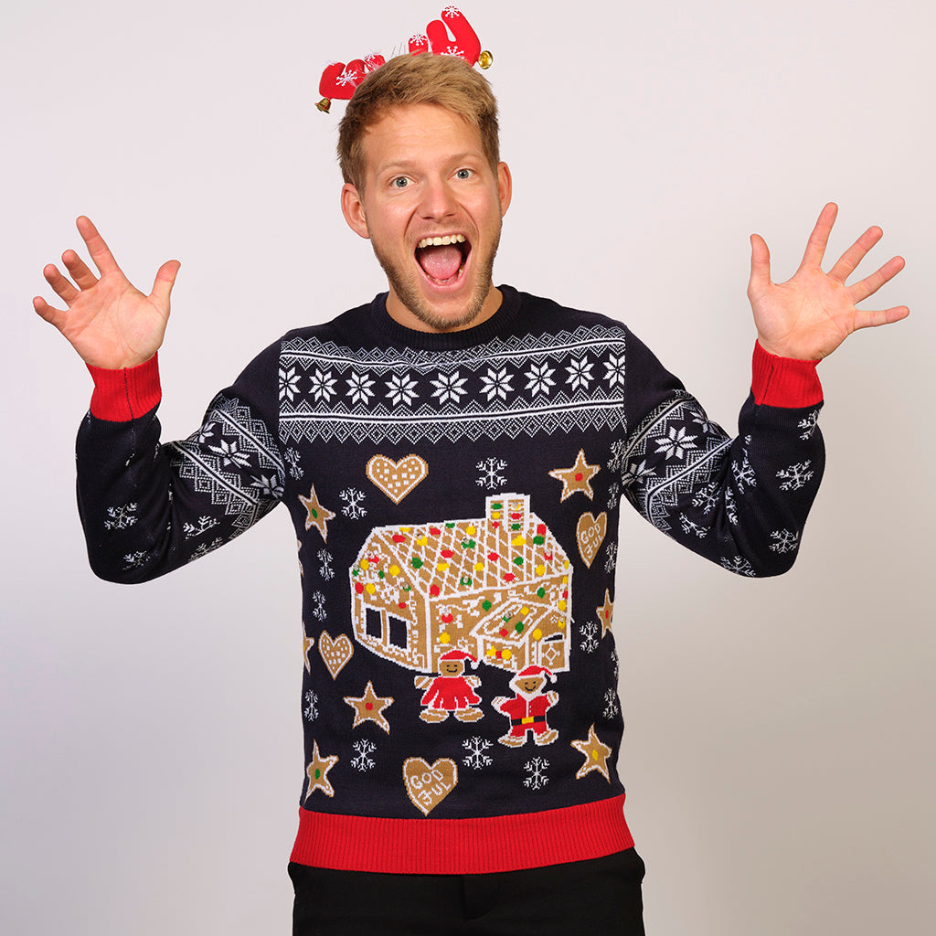 En mand poserer i en blå julesweater med et kagehus på kagemænd på.
